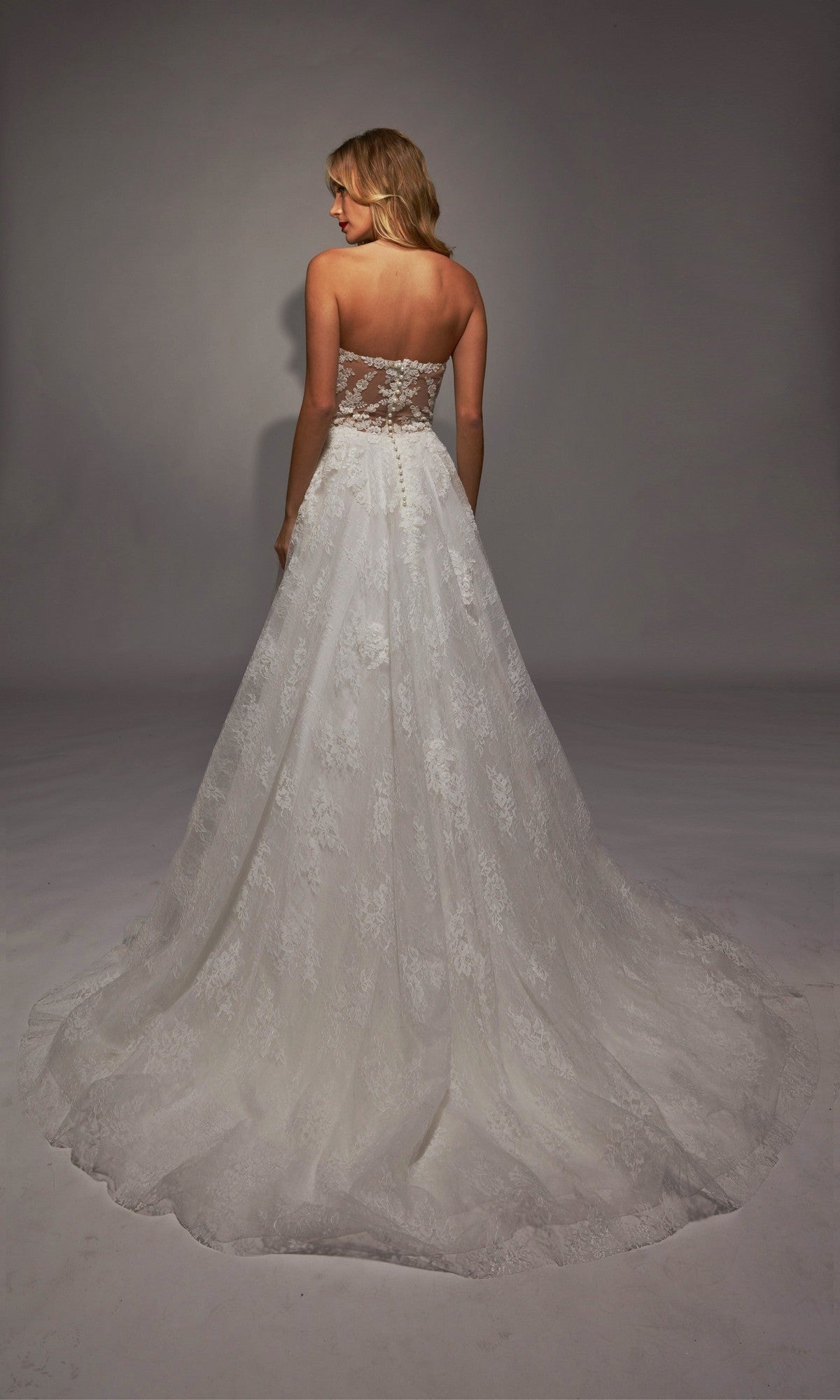 A woman wearing the Long White Dress: Alyce 7154 stands with her back to the camera, showcasing the strapless sheer corset bodice, lace ivory details, and dramatic train against a plain gray background.